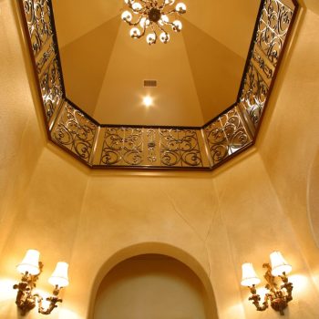 View of an ornate chandelier, decorative iron railing, and wall sconces in a domed, beige-colored entryway.