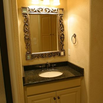 A bathroom vanity with a decorative mirror, granite countertop, sink, and three light fixtures above the mirror.