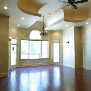 Empty living room with hardwood floors, large windows, beige walls, tray ceiling, chandeliers, and a ceiling fan.