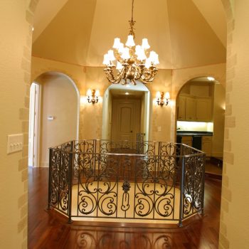 Elegant hallway with chandelier, ornate metal railing, arched doorways, and polished wood floors.