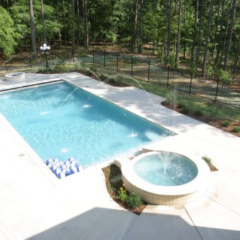 Rectangular backyard pool with water jets, a round hot tub, and blue-and-white beach balls, surrounded by trees.