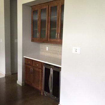 A kitchen area with brown cabinets, a built-in wine fridge, a backsplash, and white countertops.