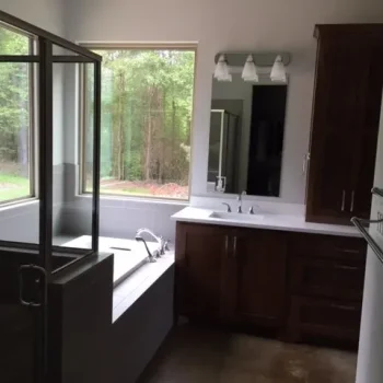 Modern bathroom with a glass shower, bathtub, wooden cabinets, and a window overlooking a wooded area.