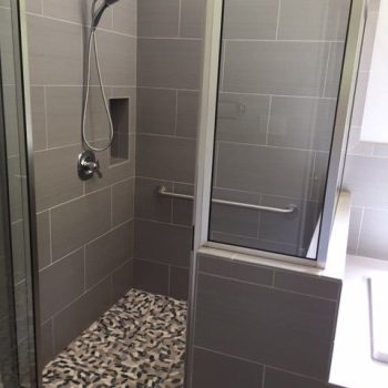 Modern walk-in shower with gray tile walls, a handheld showerhead, grab bar, and patterned tile floor.