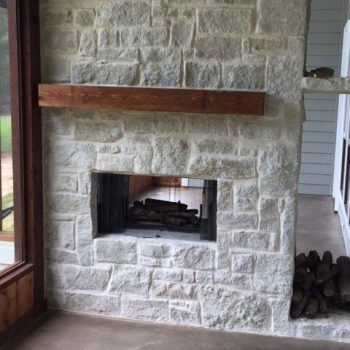 Stone fireplace with a wooden mantel, firewood storage underneath, and glass doors on a concrete floor.