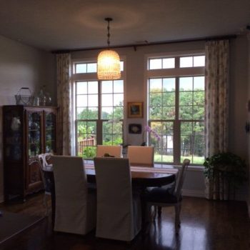 Dining room with a round table, four chairs, chandelier, large windows, and a view of greenery outside.