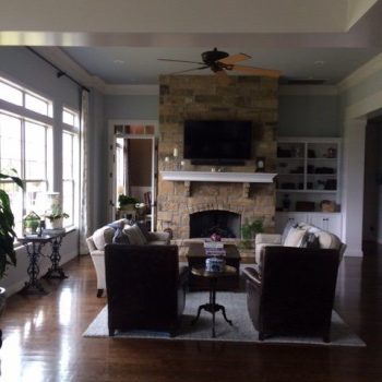 Living room with a stone fireplace, wall-mounted TV, two armchairs, and large windows letting in natural light.