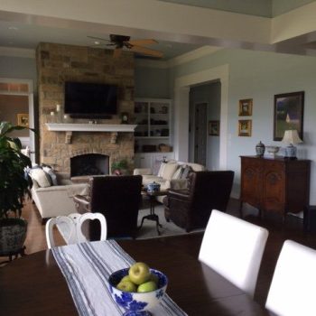 A living room with a stone fireplace, beige sofas, dark chairs, and a dining table with a bowl of fruit in the foreground.