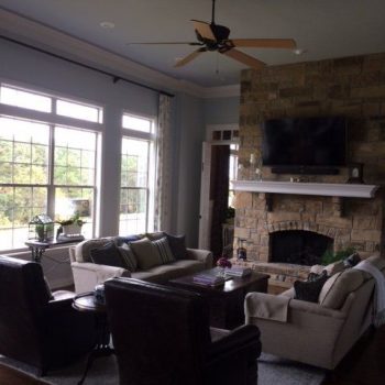 Living room with large windows, stone fireplace, wall-mounted TV, sofas, armchairs, and a ceiling fan.