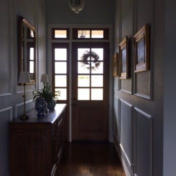 Narrow hallway with wooden floor, sideboard, framed pictures, and glass-paneled front door with wreath.