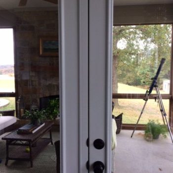 White double doors with dark handles open to a covered patio featuring a telescope, sofa, and a stone fireplace.