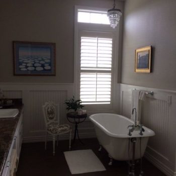 Bathroom with a clawfoot tub, a chair, a side table with a plant, two wall paintings, and a window with shutters.