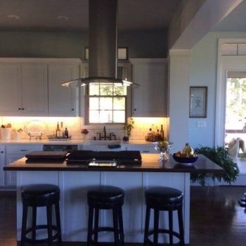 Modern kitchen with a central island, three black stools, and stainless steel range hood under a window.