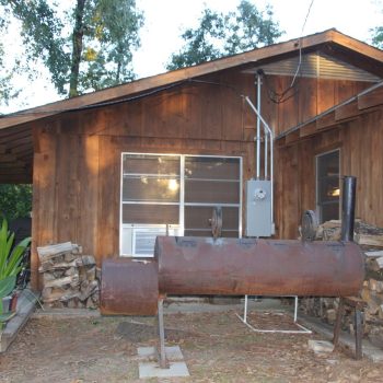 Rusty outdoor smoker grill in front of a wooden house with stacked firewood and patio chairs nearby.