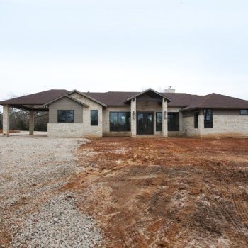 Single-story house with stone and wood exterior, brown roof, large windows, and a gravel driveway on a barren lot.