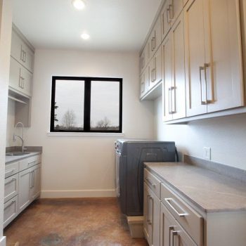 Modern laundry room with beige cabinets, gray countertops, a washing machine, and a window letting in natural light.