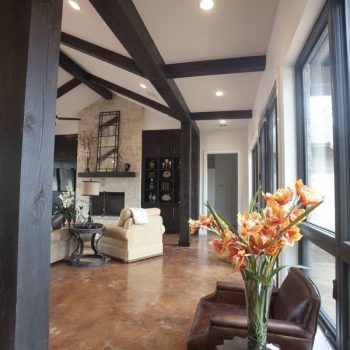 Modern living room with exposed dark wood beams, leather chairs, and orange flowers on a side table near large windows.
