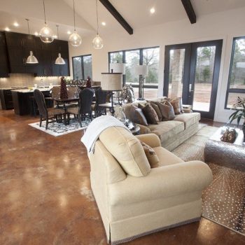Open-concept living and dining area with tan sofa, modern kitchen, large windows, and polished brown floors.