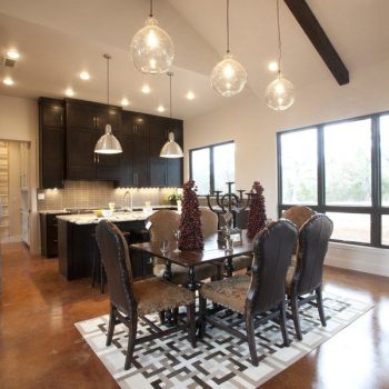 Modern dining area with six upholstered chairs, geometric rug, pendant lights, and a kitchen in the background.