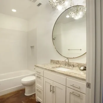 Bathroom with white vanity, round mirror, granite countertop, bathtub, and chandelier, featuring a brown floor.