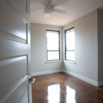 Small empty room with brown polished concrete floor, two windows, white walls, and a ceiling fan.