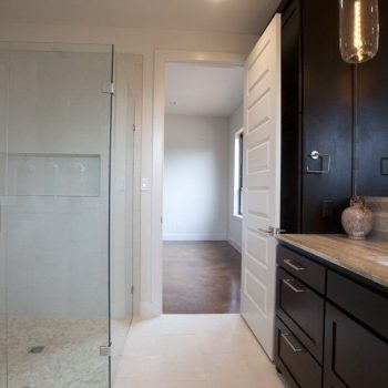 Modern bathroom with glass shower, double sink vanity, dark cabinets, and door open to adjacent room.