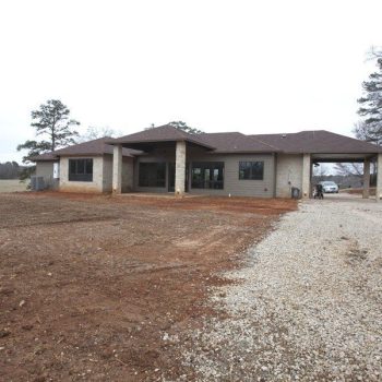 Single-story house with a covered carport, surrounded by bare soil and gravel driveway, with trees in the background.