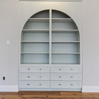 Built-in shelving unit with an arched top, four shelves, and eight drawers, against a light wall on wooden flooring.