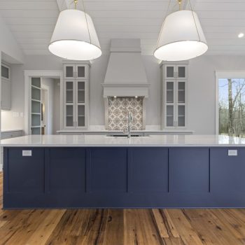 Modern kitchen with a large navy island, white countertops, pendant lights, wood floors, and large windows.