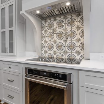 Stainless steel oven beneath a patterned tile backsplash and built-in range hood in a modern white kitchen.