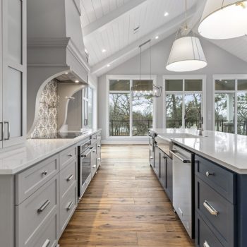 Modern kitchen with white cabinets, a blue island, wood floors, pendant lights, and large windows overlooking trees.