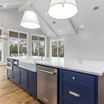 Modern kitchen with blue island, white countertops, stainless steel appliances, and large windows overlooking trees.