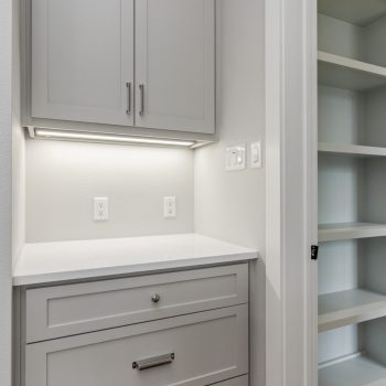 Gray built-in cabinet with drawers and shelving, white countertop, electrical outlets, and a nearby empty bookcase.