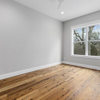 Empty room with light gray walls, wood floor, white trim, and a large window showing trees outside.