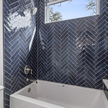 Modern bathtub with blue herringbone tile surround, chrome fixtures, and a small window above.