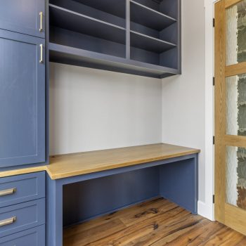 Built-in blue cabinets and desk with wood countertop, open shelving above, and a glass-paneled wooden door on the right.