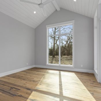 Empty room with wood floors, gray walls, large window, vaulted ceiling, and ceiling fan. Sunlight shines through window.