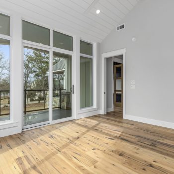 Empty room with wood floors, white walls, large windows, and a sliding glass door leading to a covered balcony with a view of trees.