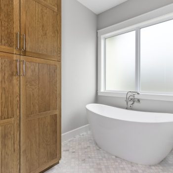 Modern bathroom with a white freestanding bathtub, large frosted window, and wooden storage cabinets.