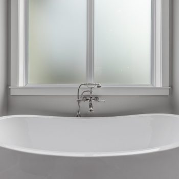 White freestanding bathtub positioned under a frosted window in a minimalist, modern bathroom.