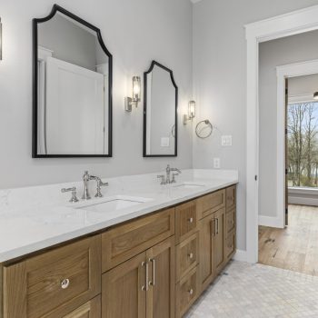 Modern bathroom with double sinks, wood cabinets, two mirrors, and wall sconces; open door shows a bright hallway.