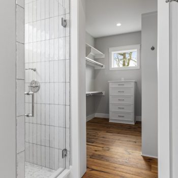 Walk-in closet with built-in shelves and drawers, next to a glass shower with white tile, and wood flooring throughout.