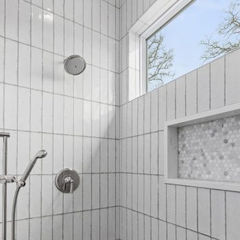 White tiled shower with two showerheads, a built-in niche, and a window showing trees outside.