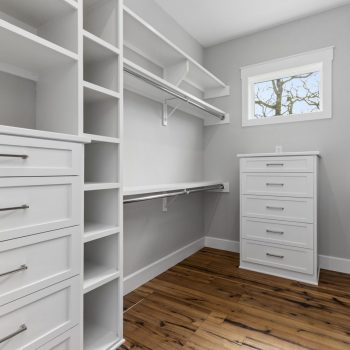 Empty walk-in closet with white shelves, drawers, and hanging rods, featuring a window and wood flooring.