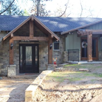 Single-story house with stone and wood exterior, black double front doors, and a small porch area, surrounded by trees.