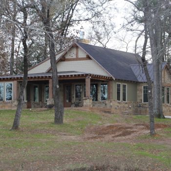Single-story house with large windows, stone and wood exterior, surrounded by leafless trees on a grassy lot.