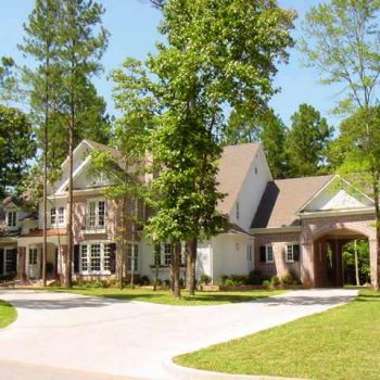 Large two-story brick house with a circular driveway, surrounded by tall trees and green lawn on a sunny day.