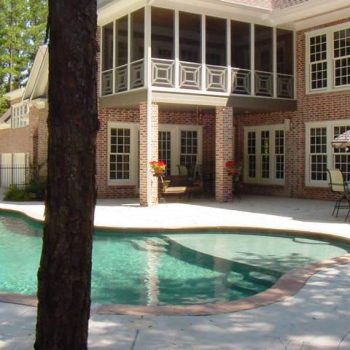 Brick house with large windows and screened porch overlooking a backyard patio and swimming pool with outdoor furniture.