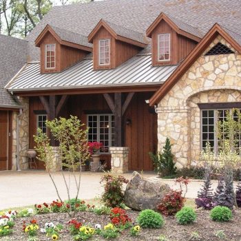 Stone and wood house with dormer windows, covered porch, and landscaped garden with flowers and shrubs in front.
