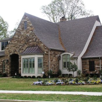 A brick house with a steep roof, arched entryway, manicured lawn, and a flower garden in front.